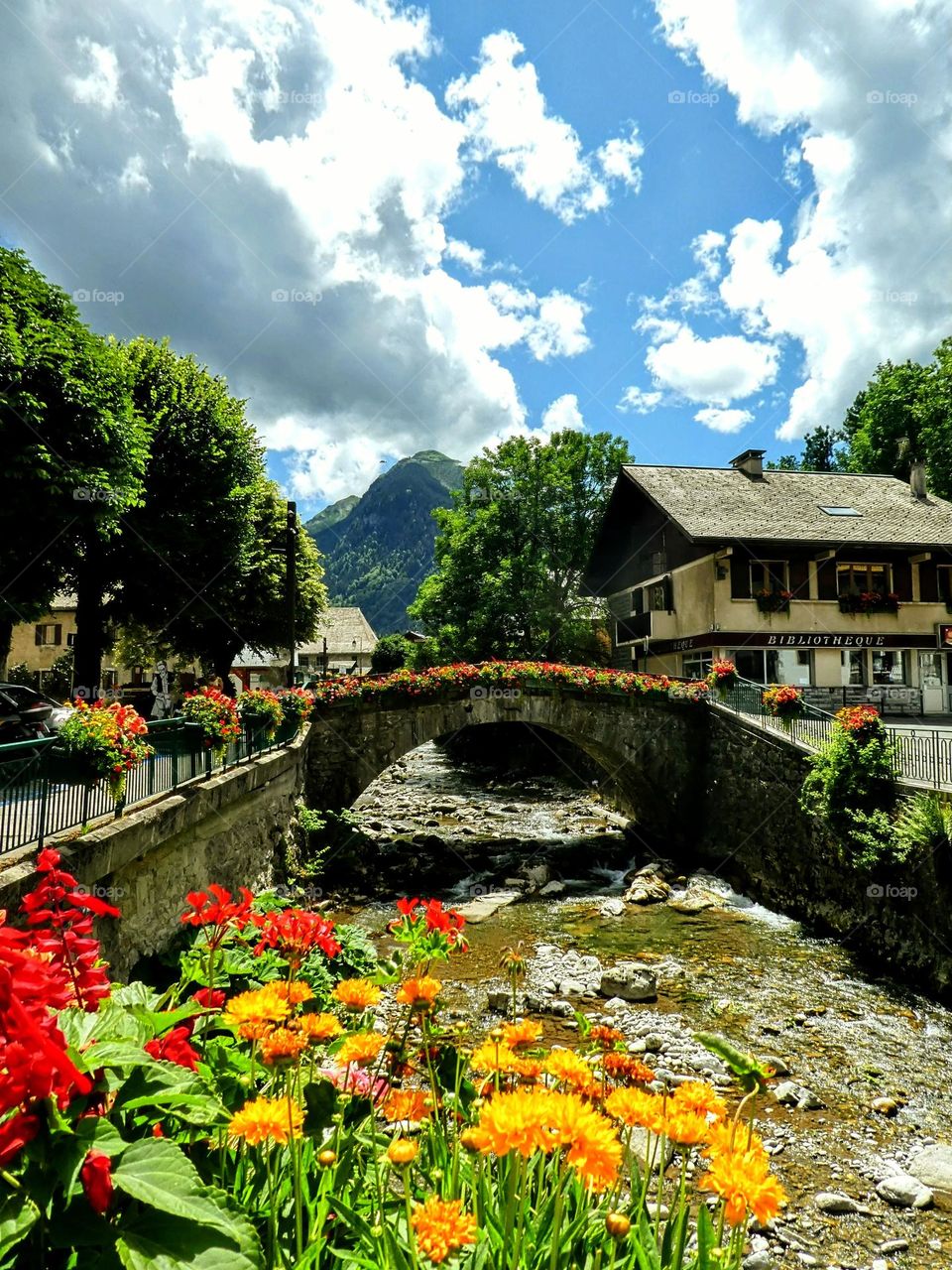 View from a bridge across the River Dranse in Morzine with red and yellow flowers in the foreground, another flowered bridge, mountains and blue sky in the background