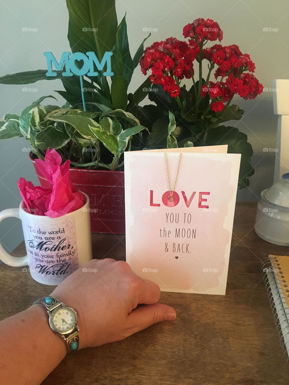 Ladies middle aged hand, wearing her favourite silver wrist watch with aquamarine stones, that she got for Mother’s day, hand curled inwards so you can only see her thumb, on a wood table, with her other gifts, flowers and card with a necklace