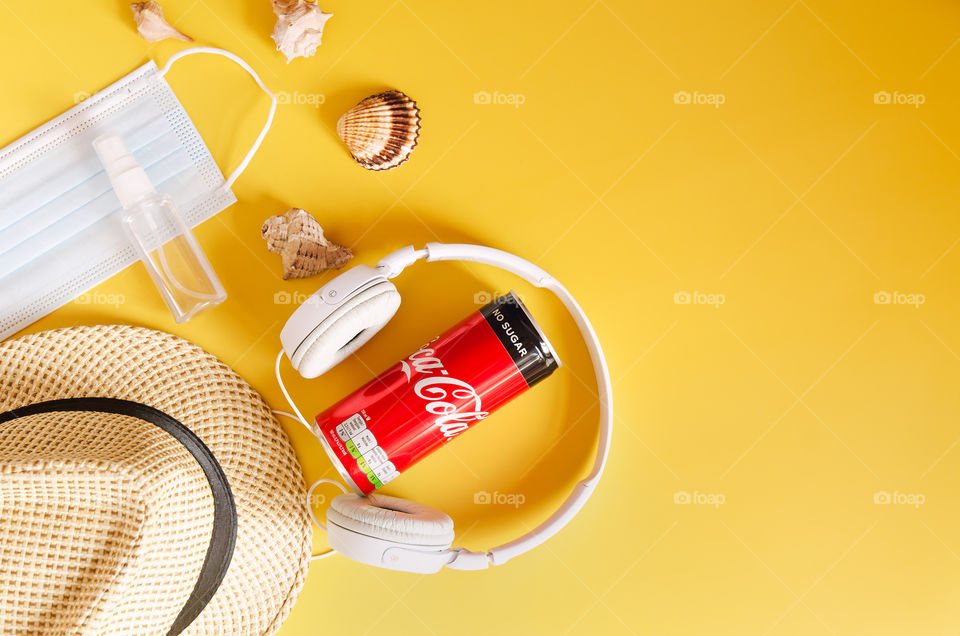 Travel under Covid-19 and new normal concept. Top view of medical face mask, can of Coca-Cola, phone and beach hat on yellow background. Creative ideas of prevent Coronavirus