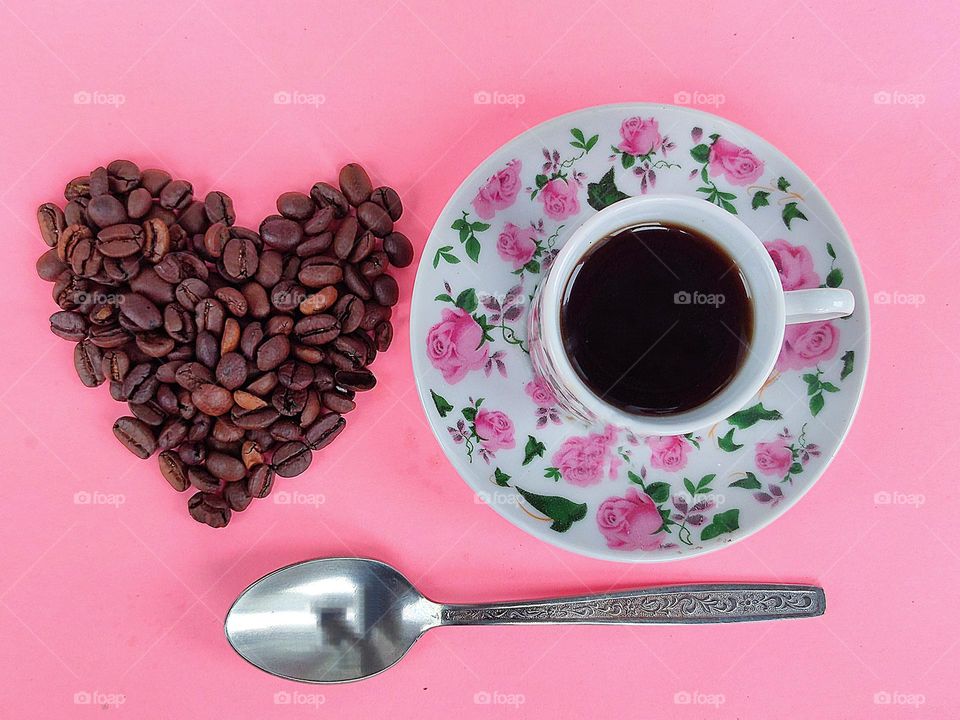 On a pink background, a saucer with pink flowers, a cup of black coffee.  Nearby on a pink background lie coffee beans in the form of a heart.  Under the heart of coffee beans and a saucer with a cup lies a silver spoon