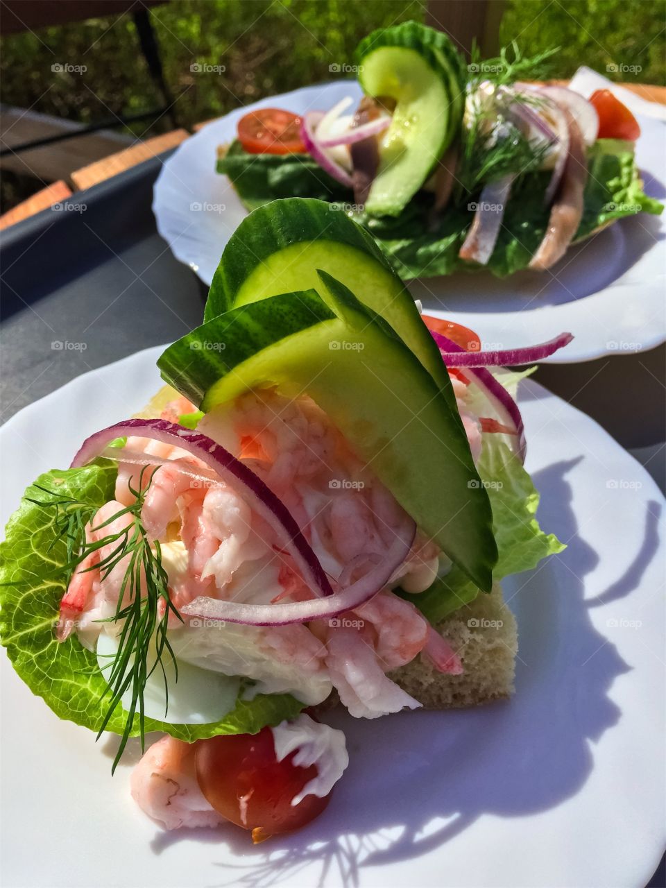 Sandwich with peeled shrimps and vegetables in foreground on white plate outdoors in garden cafe.