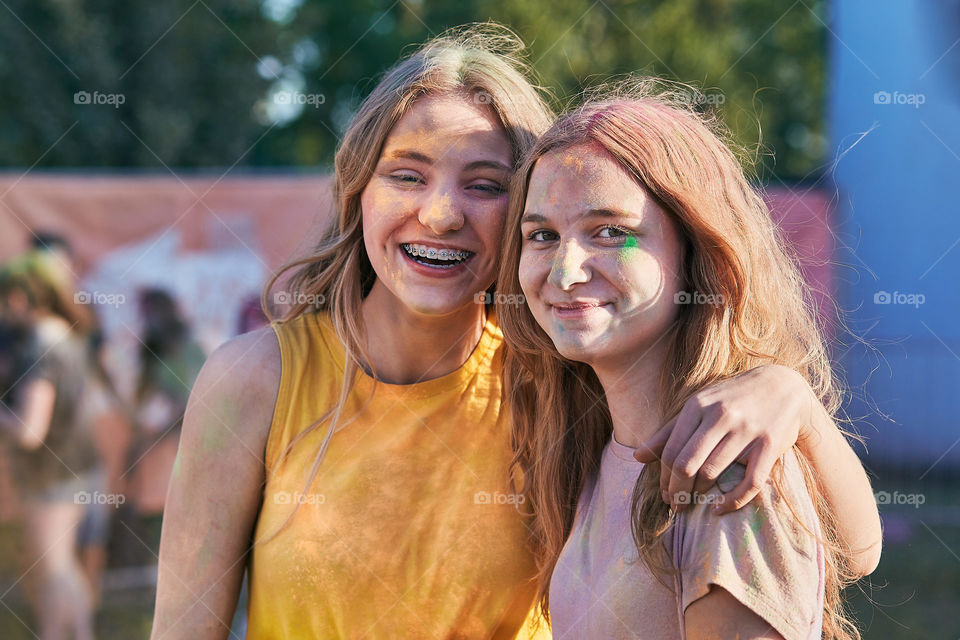 Portrait of happy smiling young girls with colorful paints on faces and clothes. Two friends spending time on holi color festival. Real people, authentic situations