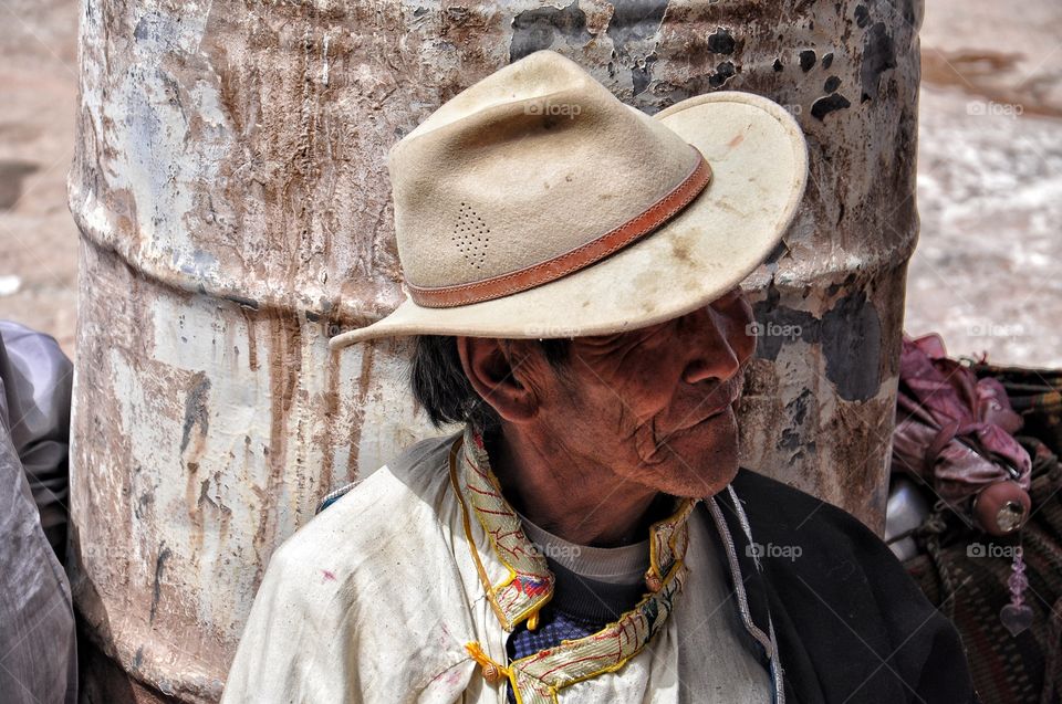 old man in the hat sitting on the ground in the yard of buddhist monastery in shigatse