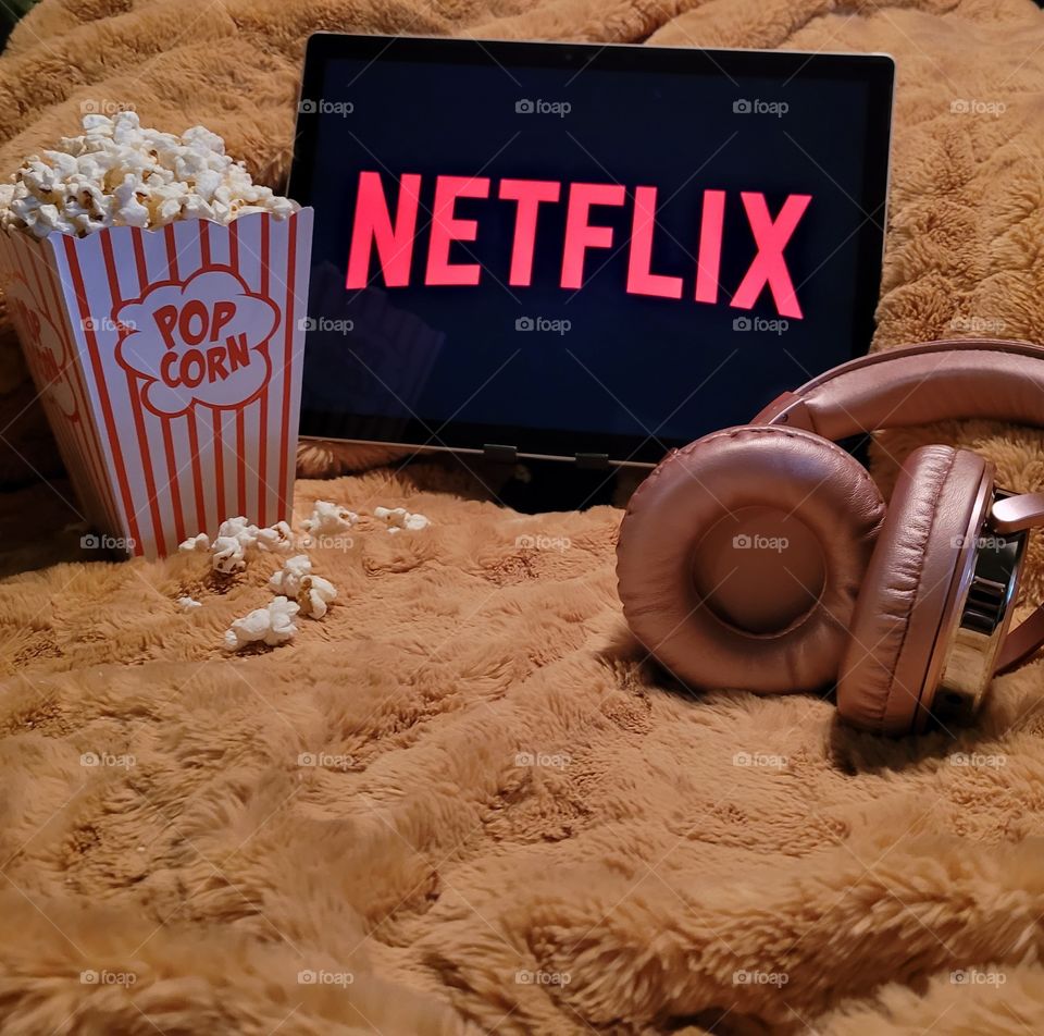 Laptop computer screen ready to watch Netflix for a fun, cozy, and relaxing night with popcorn as a snack, on soft bed blanket in room, with pink headphones