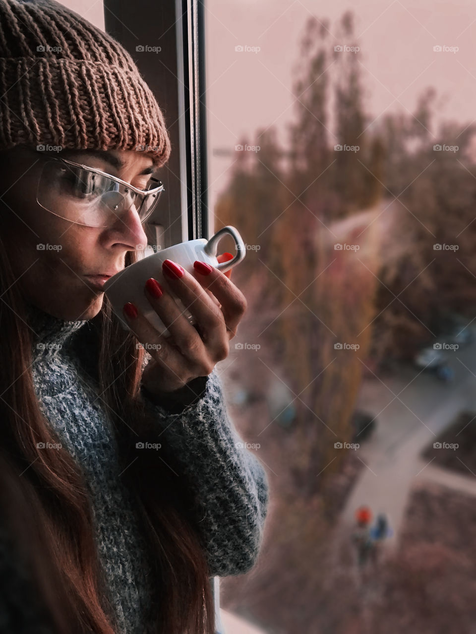 Young woman in knitted sweater and hat drinking hot coffee at the window at home 