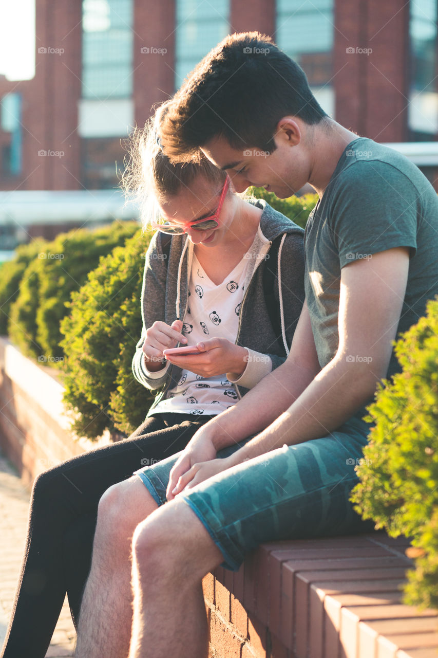 Couple of friends, teenage girl and boy,  having fun together, using smartphones,  sitting in center of town, spending time together. Real people, authentic situations