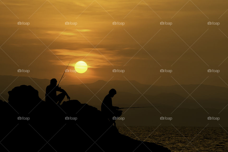 Silhouette of tourists Fishing on the rocks in sea and Golden light of sunrise behind the mountains.