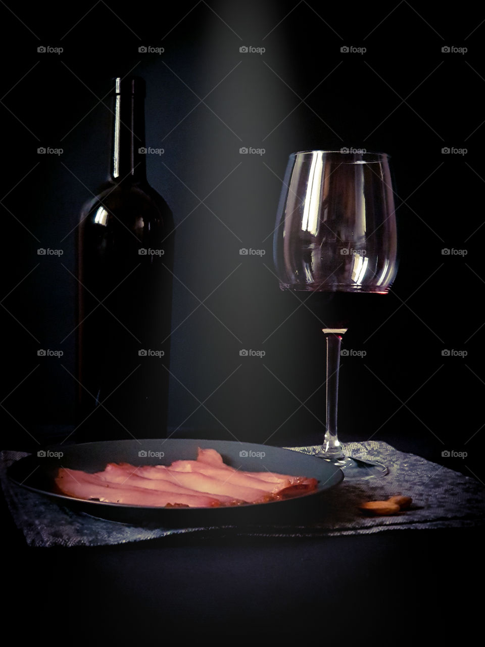 Dark still life. Black bottle of wine, a glass with red wine and a black plate with dried meat on a decorative ornamental napkin on a black background