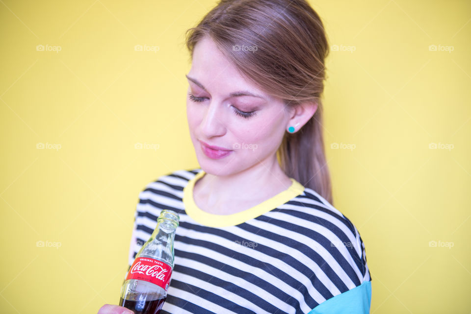 Portrait of a young woman holding a bottle of Coca-cola in front of a yellow wall