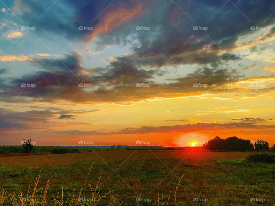 Colorful sunrise cracking the Golden sun through dark clouds under a blue sky over a farmfield and trees