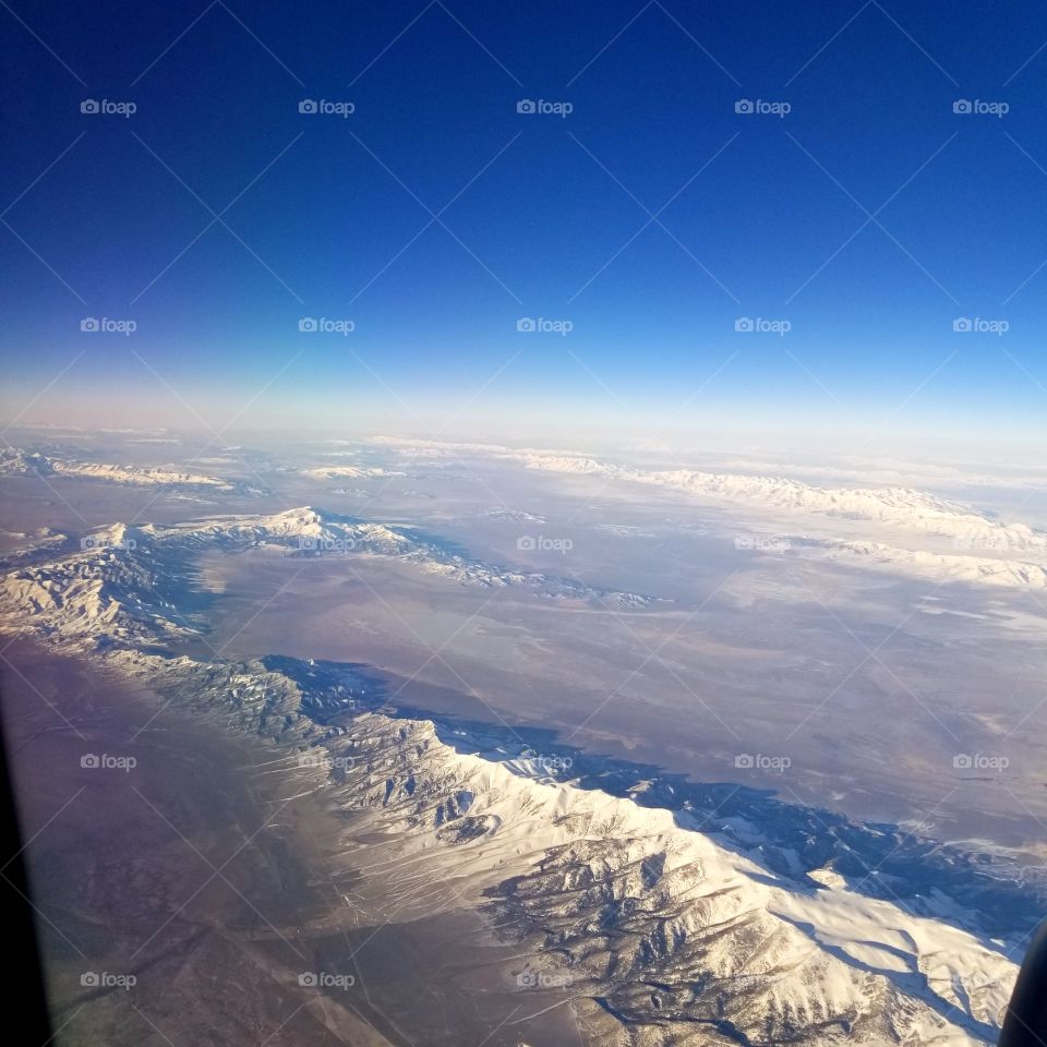 airplane view of terrain and snow covered mountain peaks with a sunny blue sky