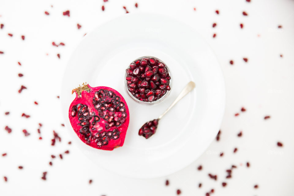 Fresh pomegranates sliced and pomegranate seeds in a glass on a white plate with single spoon. Fresh pomegranate seeds on a white background. Flat lay image of fresh healthy summer fruit.