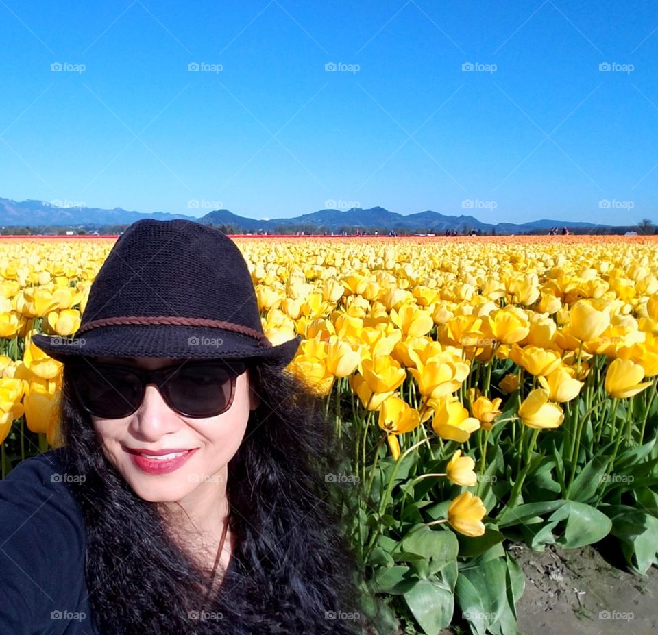A curly black hair selfie in front of a yellow tulip fields with blue sky with my sunglasses wearing a black cowboy hat and black shirt.