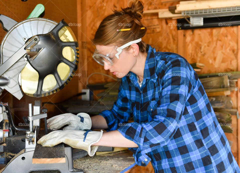 Woman cutting wood in a shed with a miter saw and wearing safety goggles and gloves