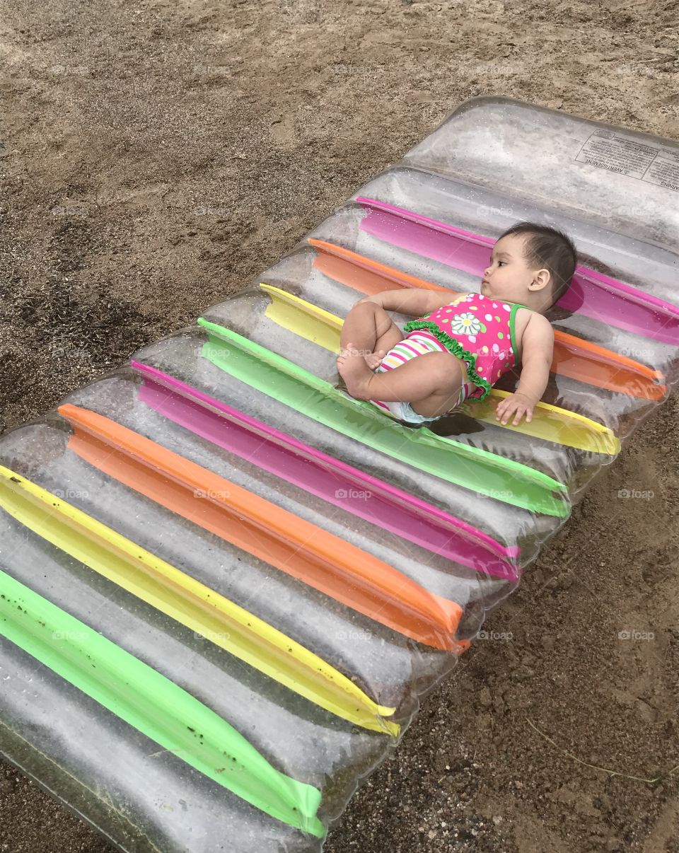 Baby girl in swimsuit on a water floating bed on sand