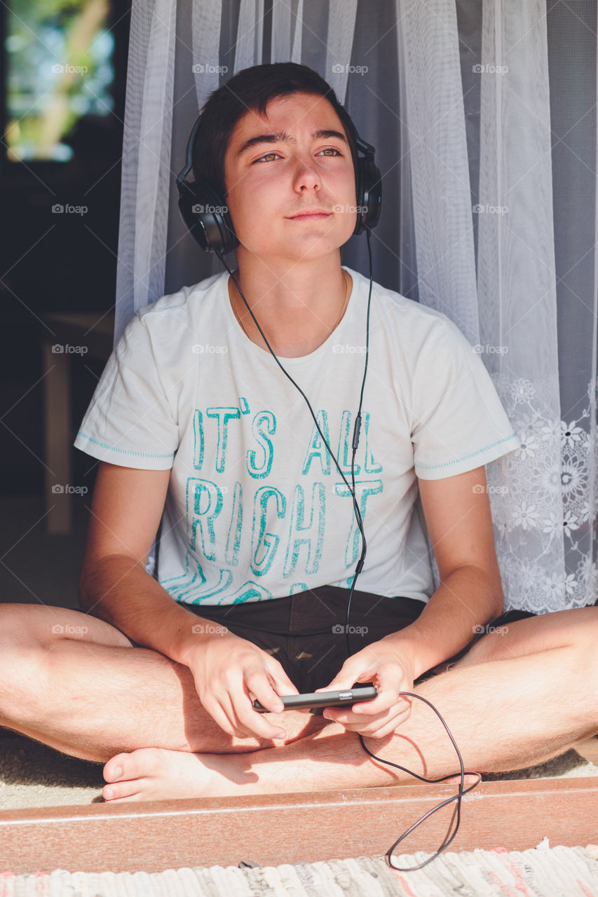 Listening to music. Young boy sitting at the patio door and listening to music from mobile phone