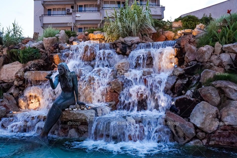 In the center of town, a mermaid statue sits on a rock in front of a waterfall. She holds a bird in her hand and listens its 🎶