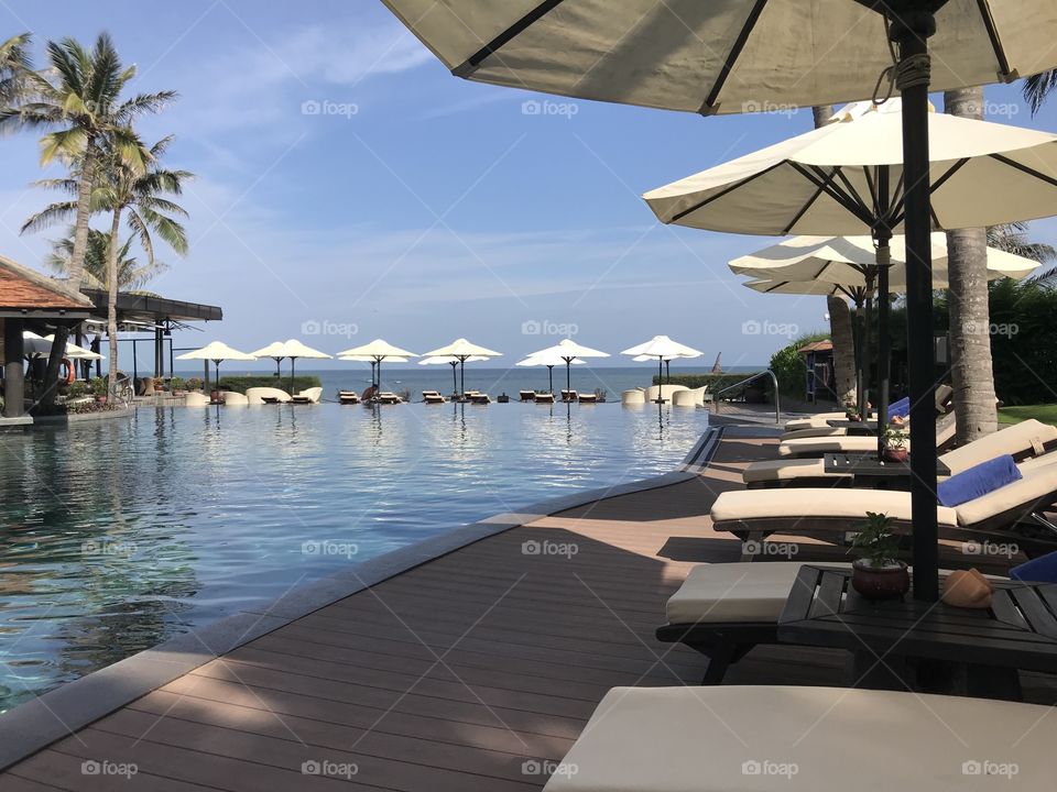 Nice swimming pool, wooden benches,white mattress,above are rolled blue towels.wooden paved outdoor walkway.there are many white umbrellas to shade the sun and a pool bar.The pool water is blue, very clean and cool.