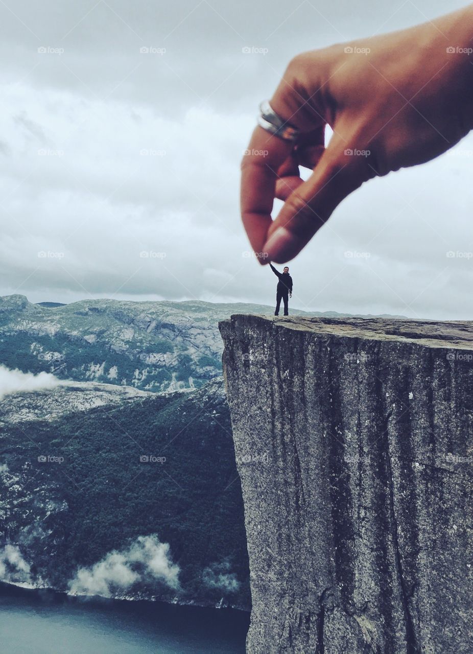 A huge hand from above is holding a tiny human silhouette standing on the edge of Prekestolen rock in Norway. 
