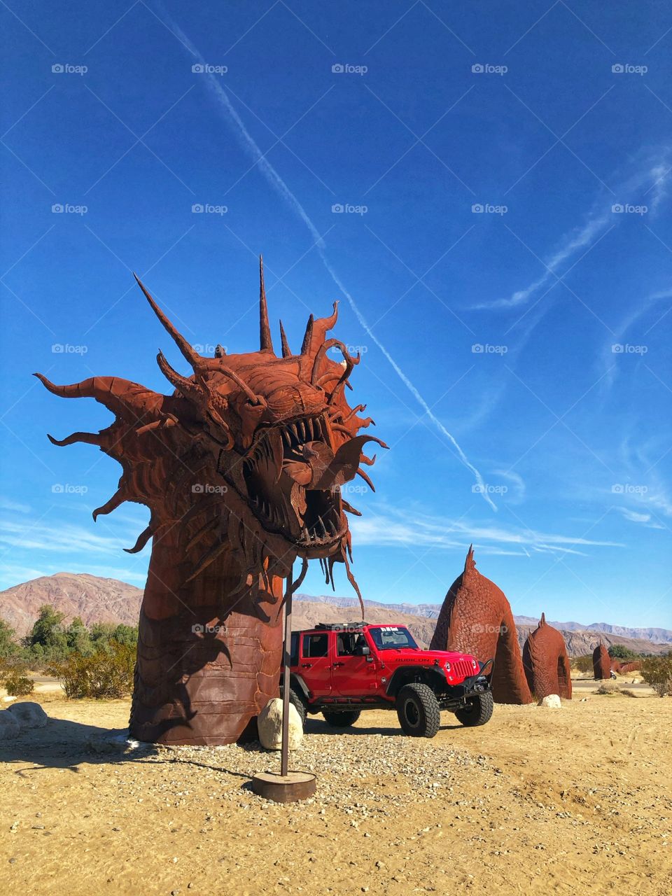Red Jeep along side large metal dragon sculpture against backdrop of big blue sky