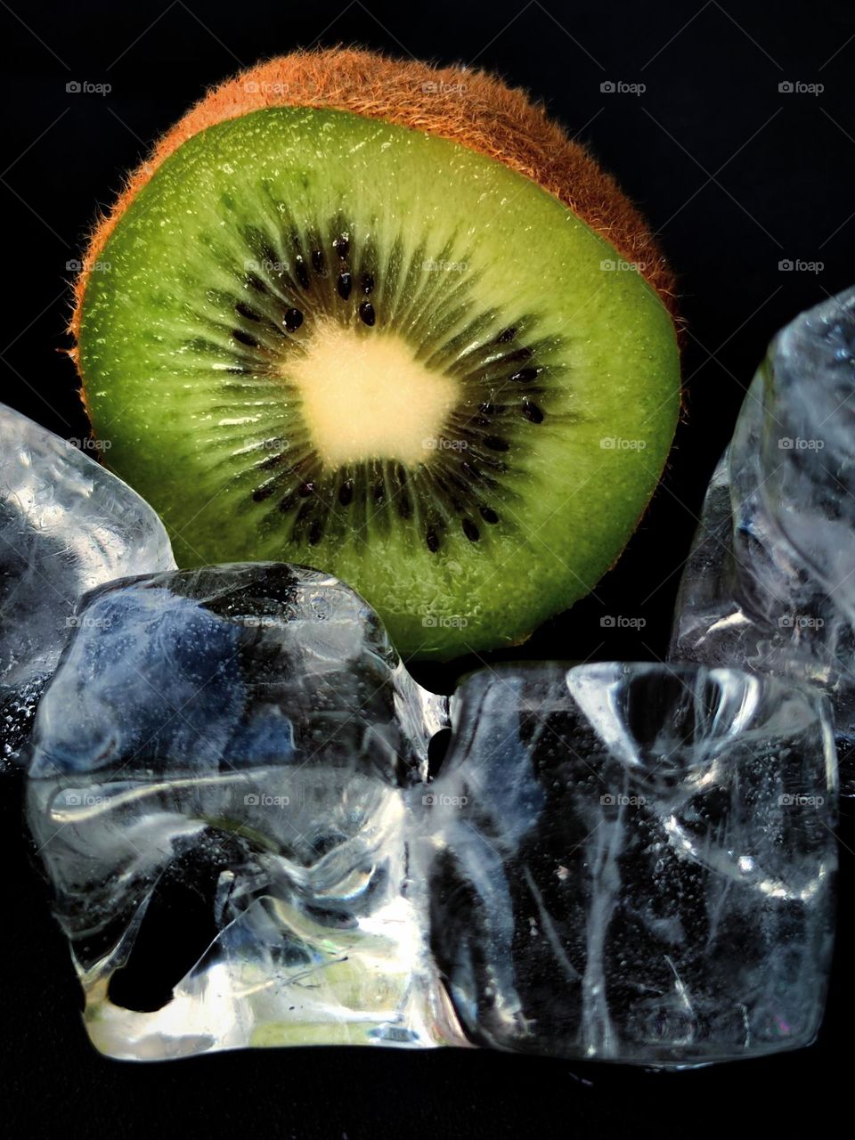 Kiwi cut.  Green flesh with black seeds.  In the foreground are ice cubes.  Juiciness contrast creates a black background