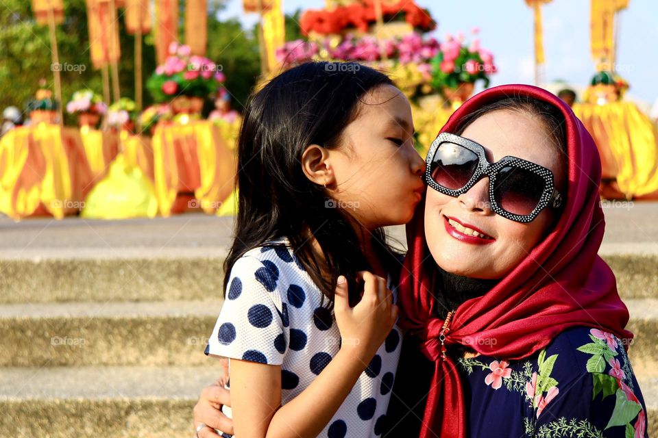 Portrait of a beautiful mother wearing a red hijab wearing a floral patterned robe and black glasses taking a photo with a happy expression being kissed by her daughter.