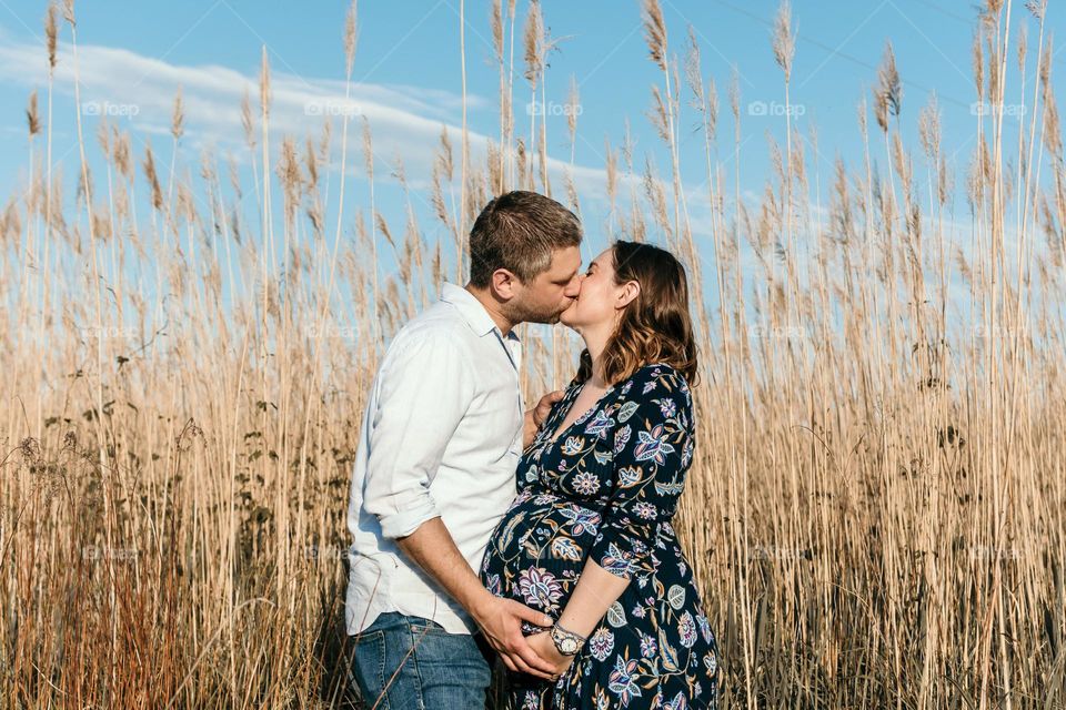 Beautiful young couple expecting a baby. Outdoor photo of happy parents to be kissing in field.