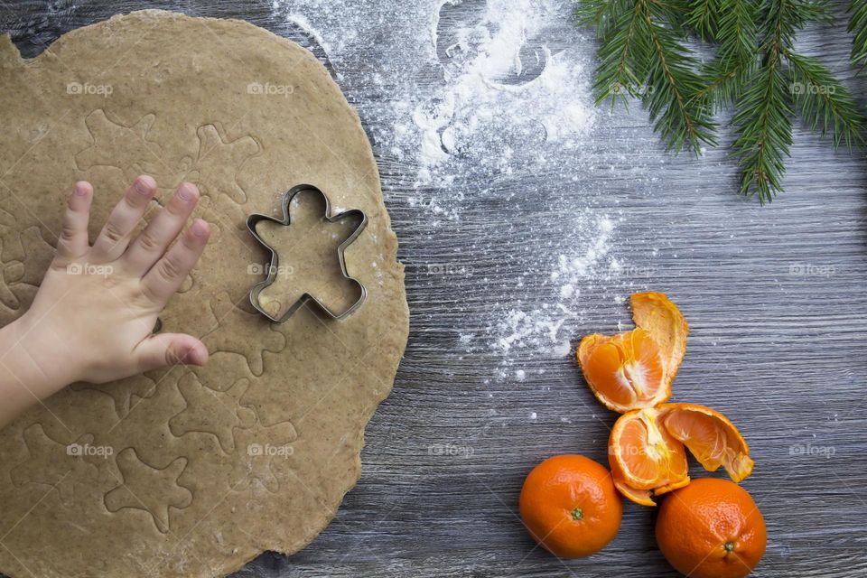 Little child helps cooking christmas ginger cookies on a wooden table with tangerines and green Christmas trees.