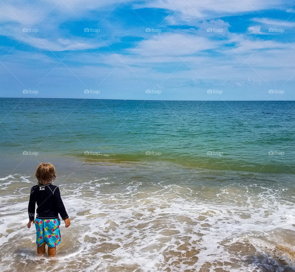 young boy standing on the beach looking out at the ocean.