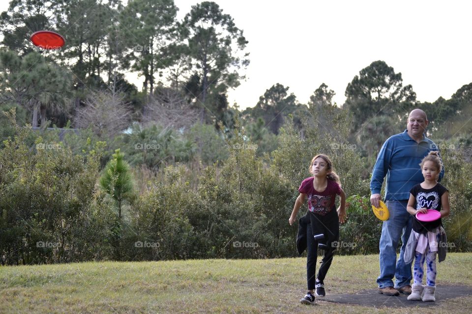 A little girl throwing a frisbee in a park with her grandfather standing beside her