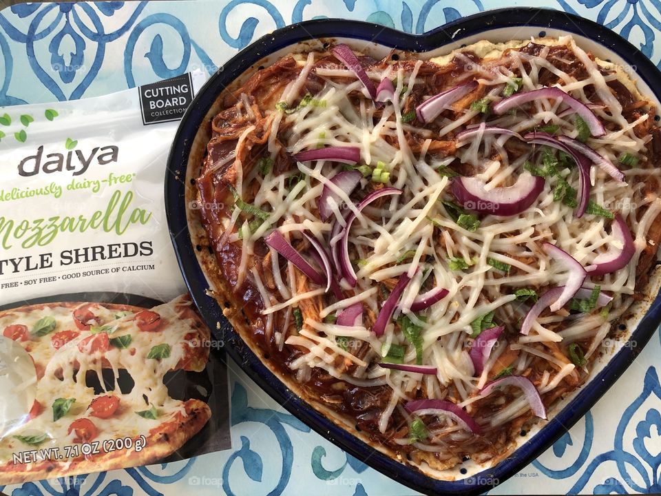 A daiya brand cheese substitute bag next to a heart shaped gluten free, dairy free, BBQ chicken pizza. 