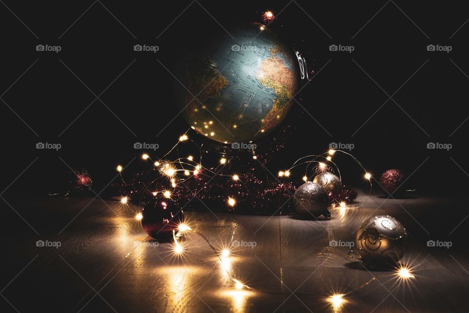 A portrait of a globe standing on a wooden floor surrounded by christmas lights and christmas balls. of depicts a worldwide christmas.