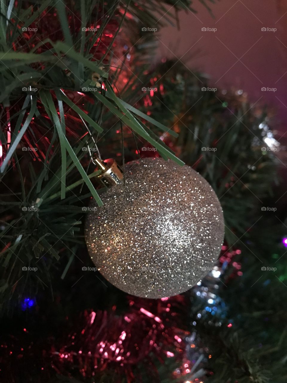 A close up of silver sparkling Christmas ornament against red tinsel and a green tree with lights around. 