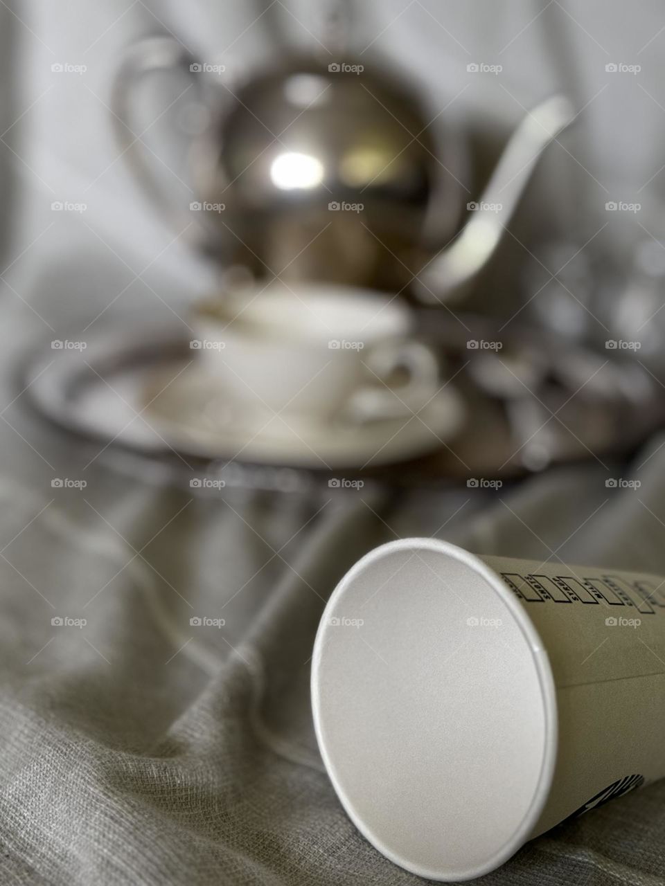 Silver coffee pot on serving dish with porcelain coffee cup and saucer with silver spoon and crystal sugar and cream set in background and paper Starbucks cup in foreground on beige woven stripe linen table cloth