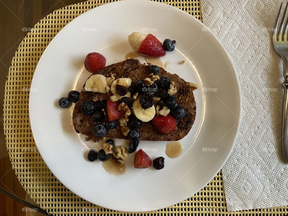 French toast with slice of strawberries, blueberries, raisins and sweet bananas for breakfast on a round white plate and yellow place mat. 