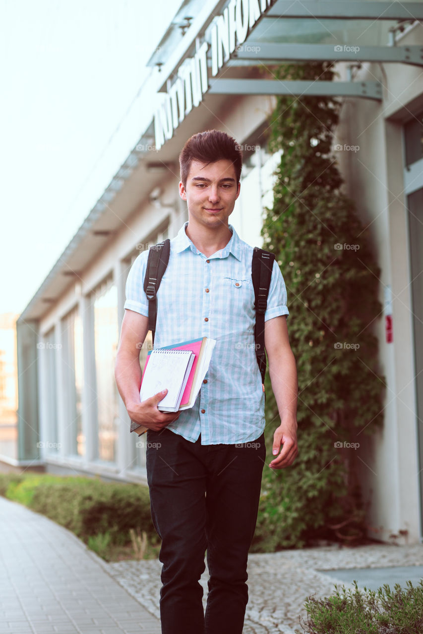 Student holding a notebook and carrying a backpack walking at the front of university building. Young smiling boy wearing blue shirt and dark jeans