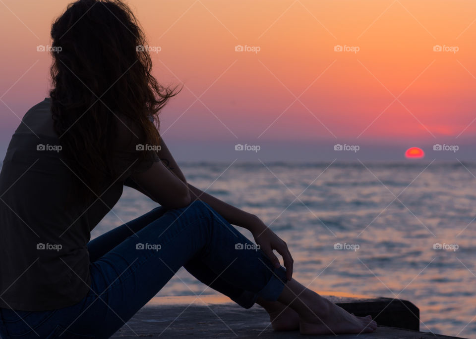 silhouette of woman sitting on a pier looking over the blue water reflecting the pinks and oranges of the sunset in the sky