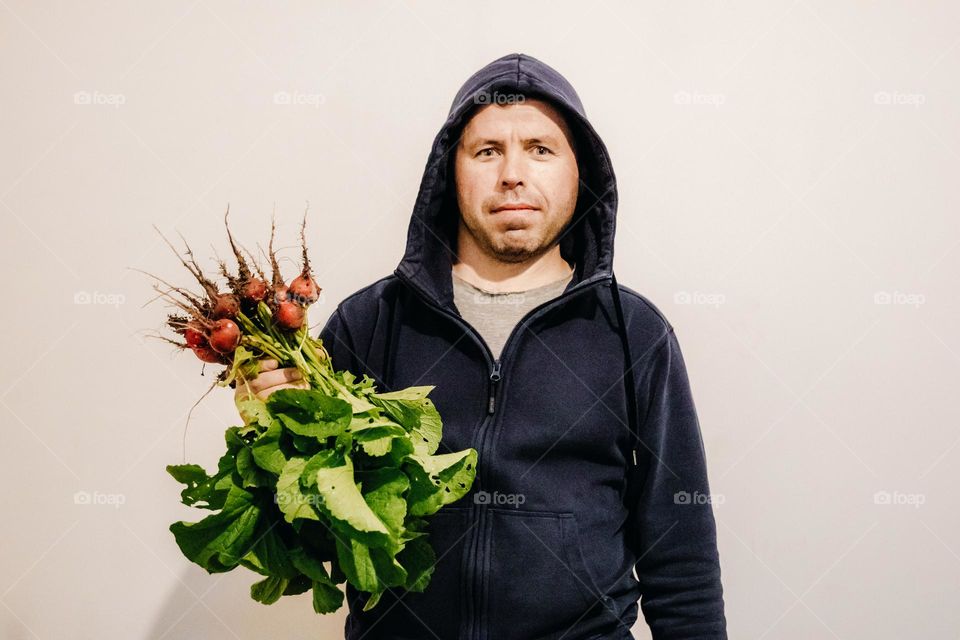 A portrait of one young handsome Caucasian man in a sweatshirt with a hoodie on his head and a bouquet of freshly picked radishes from the garden stands on a white wall in the room and looks at the camera, close-up side view.