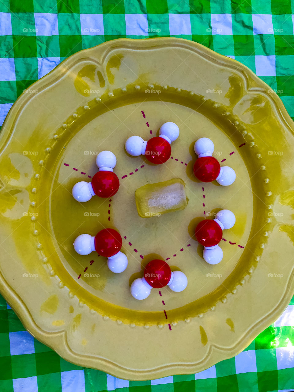 Clash of Colours: Chemistry geek alert! Red (Hydrogen) & white (Oxygen) molecule model of one ring of frozen H2O(water) surrounding an ice cube! All on a yellow plate on a green plaid tablecloth draped table. 🥶