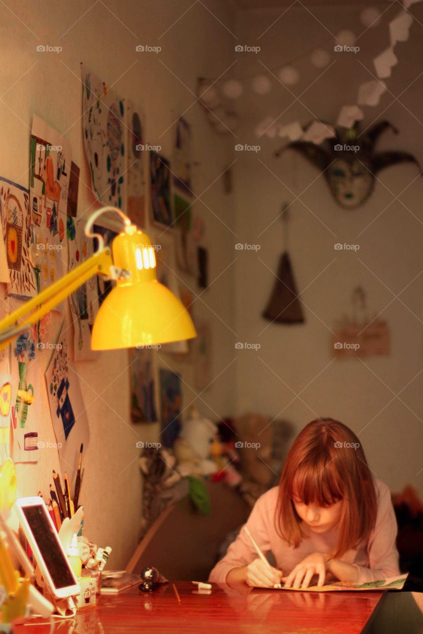 Little girl writes at a table under a yellow lamp