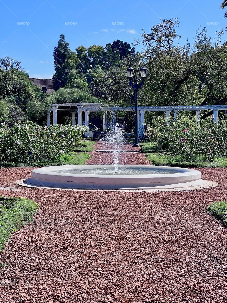 A beautiful fountain of water gushing in the center of a well-kept garden, surrounded by colorful flowers and a radiant blue sky. The path of red pebbles leads to a quiet and relaxing environment, perfect for walks.