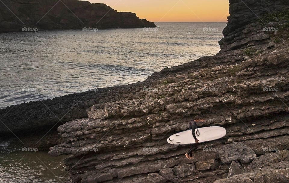 A surfer walks the rocks with his board at sunset