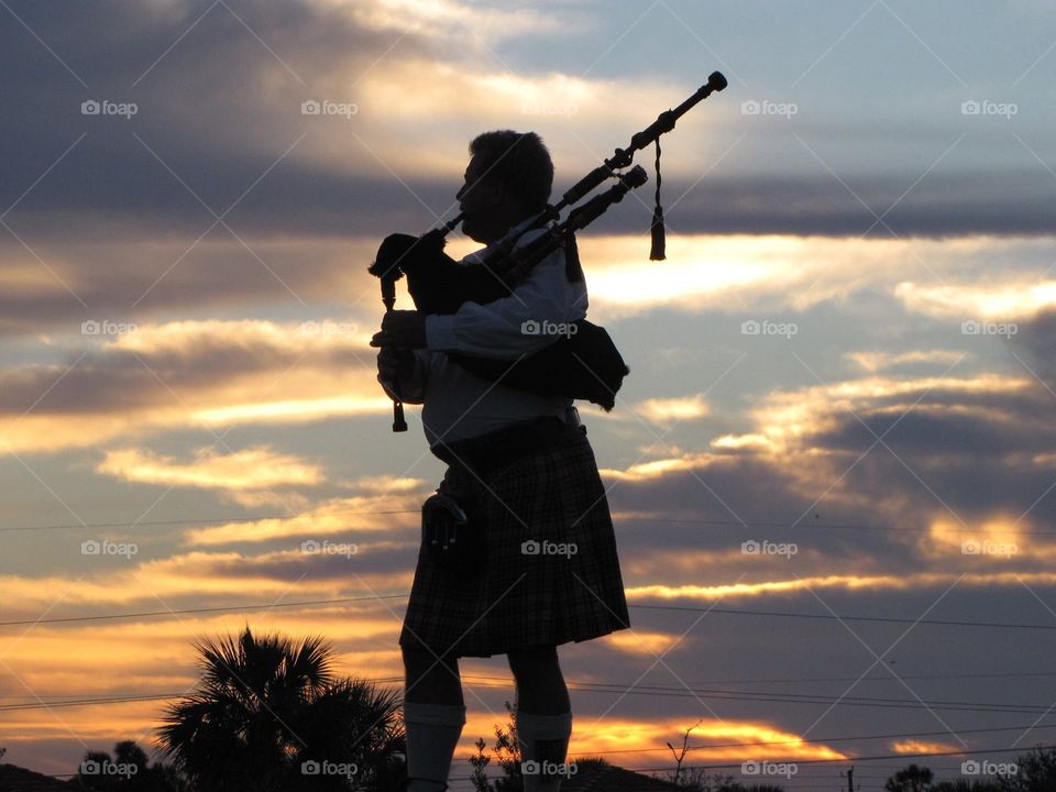 Noble, talented bagpiper playing on a hill for all to hear as the sun slowly sets behind him