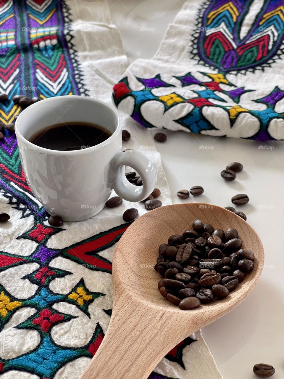 coffee theme composition: white coffee cup containing liquid coffee and coffee beans on white surface and inside a natural wooden spoon, on hand-embroidered Eritrean drape with typical ethnic embroideries