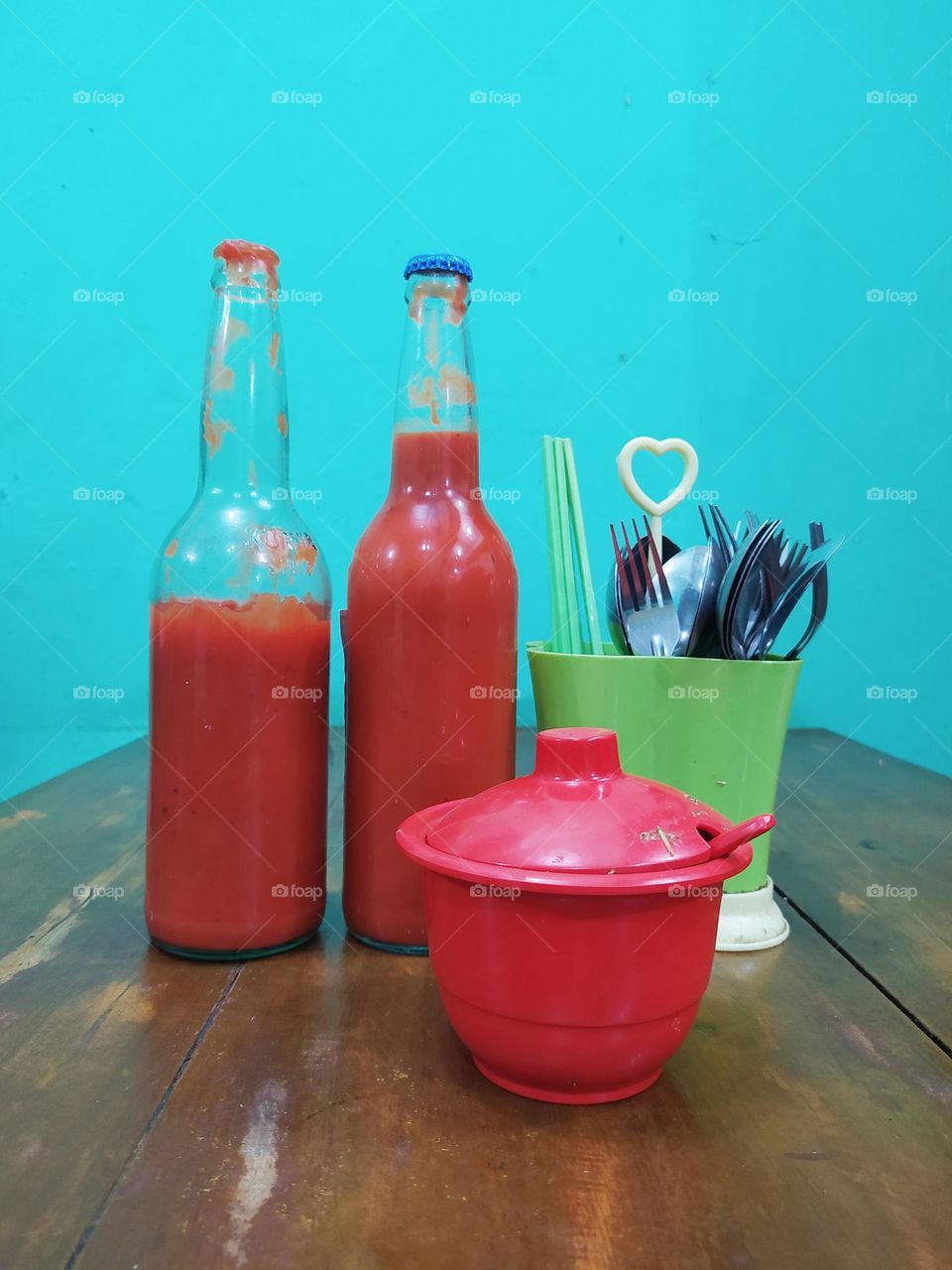 View of the dining table at a local chicken noodle shop, there are bottles of tomato sauce, chili sauce, cutlery, chopsticks on a wooden table with a Tosca wall as a background