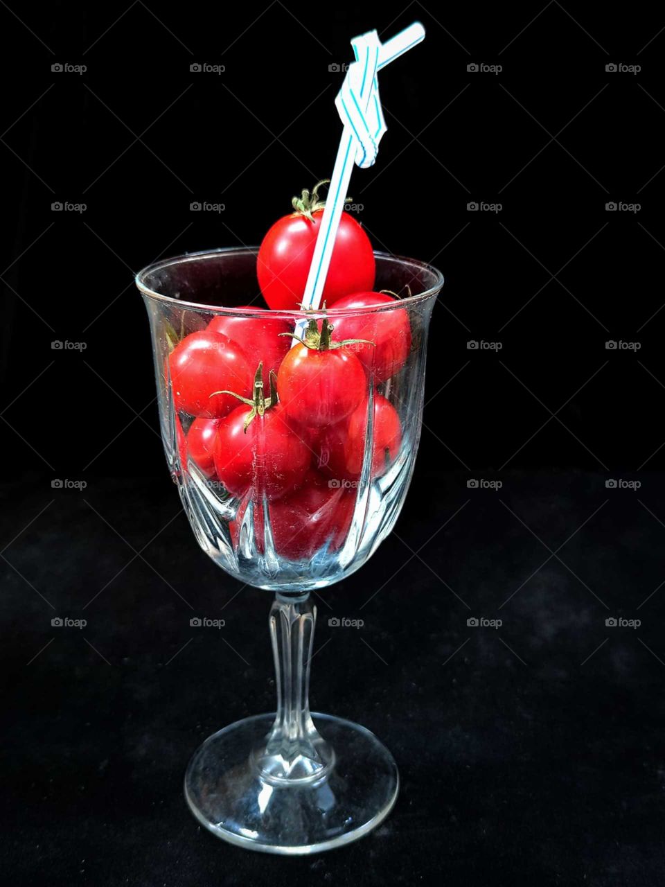 Wine glass with red cherry tomatoes and a blue  tube, the end of which is tied with a knot.  Black background