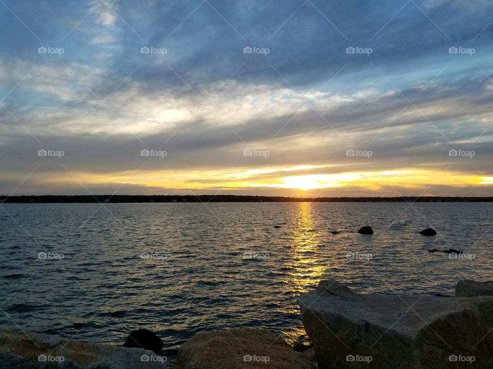 Sunrise, Sunsets in USA! Sun low on horizon, lighting clouds & showing reflection lighting up shores' rocks near inlet in New England.