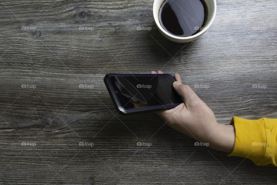 A man drinks coffee in the morning and works on the phone. Men's hands hold a white cup of coffee and a mobile phone in a yellow jacket. The hands lie on a gray wooden surface that is used as a background.