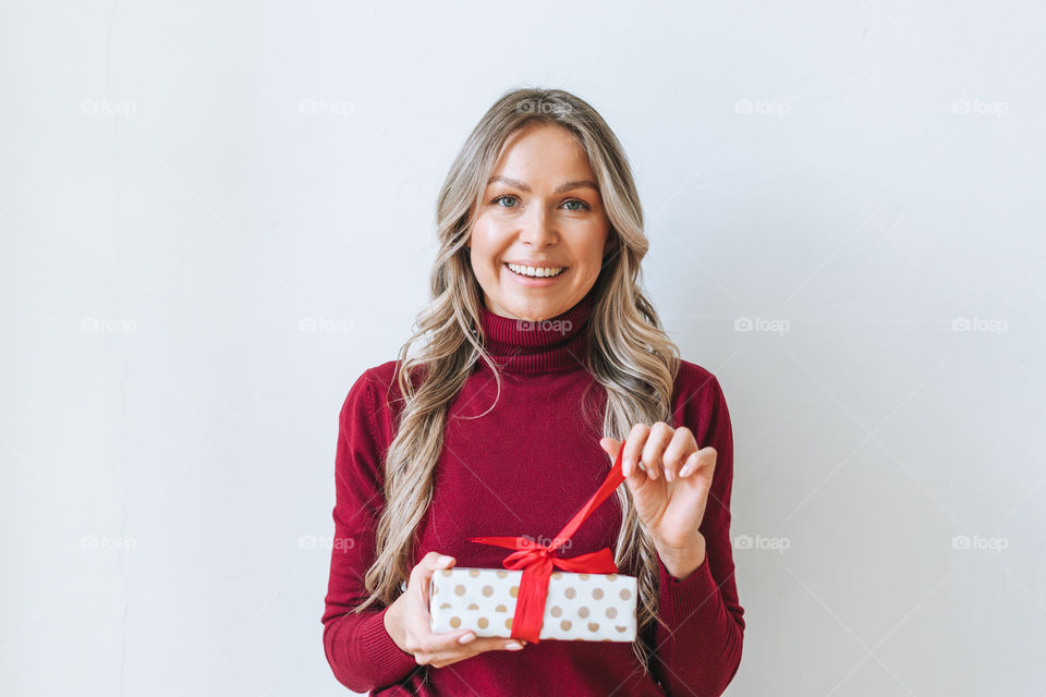 Young blonde woman in bordo sweater holding gift box present in hands on grey background 