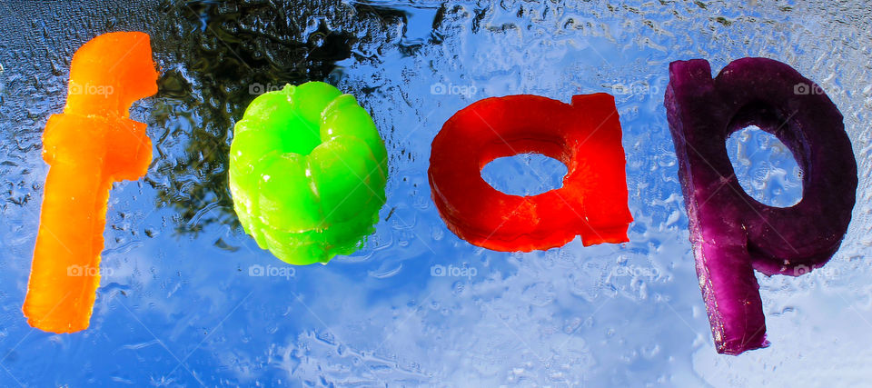 The foap name spelled out in jello letters and placed on a mirror outside on a sunny, partially cloudy, day.  There is a reflection of the clouds and a tip of a tree. The mirror was misted with water making the letters seem they were also in water. 